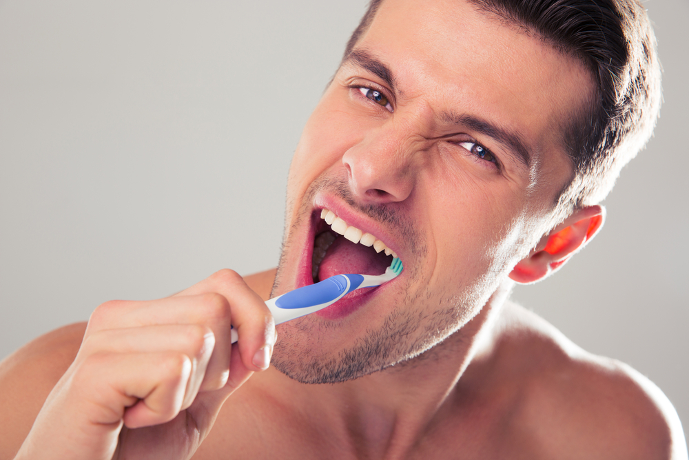 Handsome man brushing teeth over gray background. Looking at camera